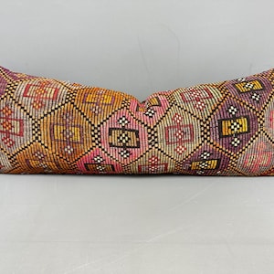 Op de afbeelding: Een lang, rechthoekig kussen met een kleurrijk, geometrisch patroon. Het kussen heeft een mix van warme kleuren, waaronder oranje, roze en geel, met accenten van paars en rood. Het ontwerp is ingewikkeld en gedetailleerd, waardoor een textuur ontstaat.