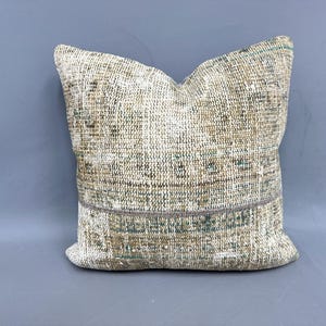May include: Square decorative pillow with a textured, woven design. The pillow features a mix of neutral colors, including beige, brown, and hints of green. A darker band runs across the middle, adding a contrasting element. The pillow is a home decor item.
