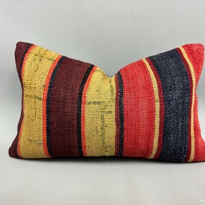 May include: Rectangular pillow with a striped pattern in shades of red, gold, and navy blue. The pillow has a textured appearance, suggesting a woven fabric. The design features vertical stripes, creating a visually appealing accent piece.