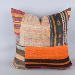 May include: Square decorative pillow with a patchwork design. The pillow features a variety of colors, including orange, red, black, and beige. The pillow is made of a textured fabric and is likely used for home decor.