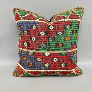 May include: Square pillow with a woven, geometric pattern in red, green, blue, and white. The pillow has a textured appearance and a decorative border. The design features repeating squares and diamond shapes, creating a vibrant, colorful accent.