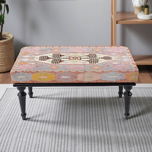 May include: A rectangular ottoman with a patterned top in shades of pink, gray, and yellow. The design resembles a traditional rug. The ottoman has a black frame and four black, turned legs. The piece is placed on a gray rug.