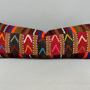 May include: A rectangular pillow with a patchwork design. The pillow features a central pattern of red, black, and brown geometric shapes, bordered by stripes of orange, blue, and brown. The pillow is a decorative item.