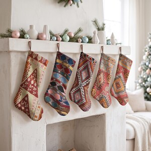 Puede incluir: Cinco calcetines de Navidad colgados en fila de una repisa de chimenea blanca. Cada calcetín presenta un patrón tejido distintivo y colorido con formas geométricas, incluyendo tonos de rojo, azul, verde y naranja, suspendidos por lazos marrones.