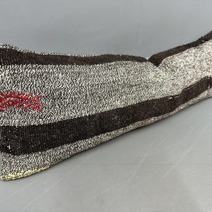 May include: A rectangular pillow with a textured, woven appearance. The pillow features alternating bands of dark brown and light gray. A small red embroidered design is visible on one end. The pillow is designed for decorative use.