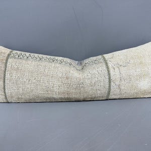 May include: A long, rectangular pillow with a textured, off-white fabric. The pillow has a faded appearance with a darker stripe down the center and at each end. The pillow is likely for home decor.