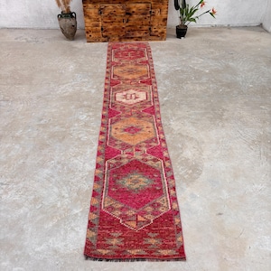 May include: A long, narrow runner rug featuring a vibrant magenta and deep red base color. The rug displays a repeating pattern of large, interconnected hexagonal medallions in shades of orange, cream, and muted green, finished with short fringe ends.