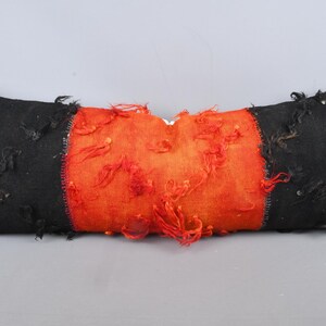 May include: A rectangular decorative pillow with a black and orange color scheme. The pillow has black ends and an orange center section. The orange section has red, textured accents. The pillow is designed for home decor.