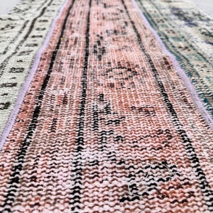 Peut inclure: Gros plan sur un tapis tissé rose et blanc avec des motifs géométriques noirs et bruns. Le tapis a des franges aux deux extrémités.