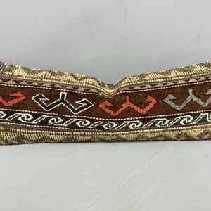 May include: Rectangular decorative pillow with a brown and beige woven design. The pillow features a central band of dark brown with geometric patterns in orange, white, and gray. The edges have a lighter, textured pattern.