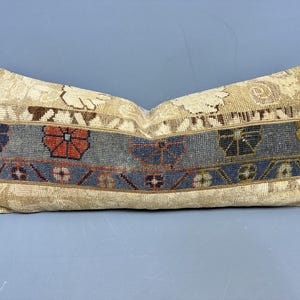 Peut inclure: Coussin rectangulaire décoratif avec un fond beige et une bande centrale bleu-gris avec des motifs floraux et géométriques rouges, dorés et bleus. Le coussin a un aspect vintage et texturé.