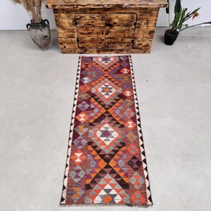 May include: A rectangular, patterned rug with a geometric design. The rug features a central diamond pattern in shades of orange, red, purple, and gray. The edges are bordered with a black and white zigzag pattern. The rug is placed on a light gray surface.