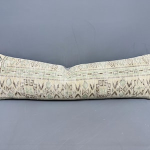 May include: Rectangular decorative pillow with a cream-colored background and a repeating geometric pattern in shades of brown and green. The pillow is long and narrow, suitable for a sofa or bed.