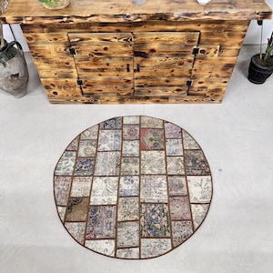 May include: A circular patchwork rug with a brown border. The rug features a variety of rectangular and square fabric pieces in muted tones such as beige, blue, and red. It is placed on a light grey surface.