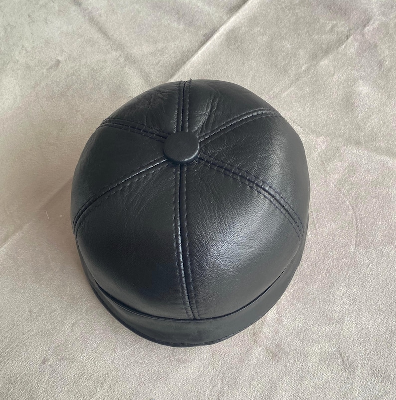 May include: A black leather cap with a round crown and a stitched design. The cap has a button on the top.
