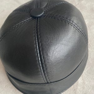 May include: A black leather cap with a rounded crown and a flat brim. The cap has a button on top and is stitched with a contrasting thread.