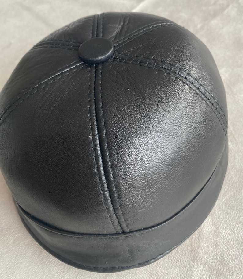 May include: A black leather cap with a rounded crown and a flat brim. The cap has a button on top and is stitched with a contrasting thread.