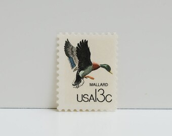 May include: A vintage US postage stamp featuring a mallard duck in flight. The stamp is white with black and green accents and reads "USA 13c" and "Mallard".