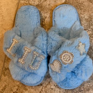 May include: A pair of light blue faux fur slippers with a crisscross strap design. The left slipper features the letters L, I, and U in white pearl-encrusted block letters. The right slipper features a pearl-encrusted heart, flower, and star.