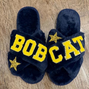 May include: A pair of navy blue, fluffy cross-band slippers. The slippers feature the word "BOBCAT" in yellow block letters, with a gold glitter star on each slipper. The slippers are on a wooden surface.