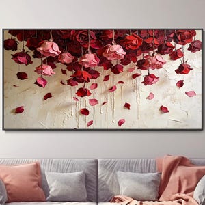 Puede incluir: Una obra de arte en lienzo enmarcada con un fondo texturizado con rosas rojas y rosas colgantes y pétalos dispersos. La obra de arte cuelga sobre un sofá gris con cojines rosas y grises y una manta rosa.