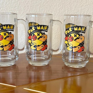 May include: Three clear glass beer mugs with handles. Each mug features a colorful Pac-Man graphic with the words "PAC-MAN" in a black rectangle. The Pac-Man character is yellow, with red and orange ghosts.
