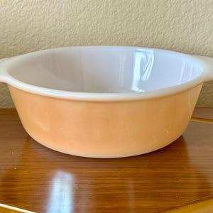 May include: An orange and white Pyrex bowl with two handles. The bowl has a rounded shape and is sitting on a wooden surface. The interior of the bowl is white, and the exterior is a solid orange color.