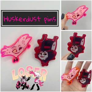 Könnte beinhalten: Zwei pinkfarbene und rote Emaille-Pins mit Comicfiguren aus der Serie "Hazbin Hotel". Die Figuren sind Angel Dust und Alastor. Die Pins befinden sich auf einem weißen Hintergrund mit dem Text "Huskerdust pins" darüber.