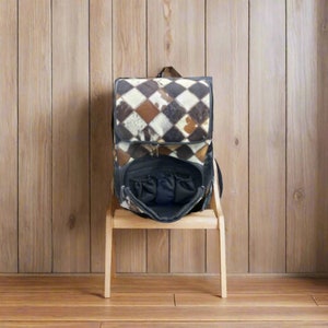 May include: A black leather backpack with a patchwork design of brown, white, and black cowhide. The backpack is open and the inside is lined with black fabric. The backpack is sitting on a wooden stool.
