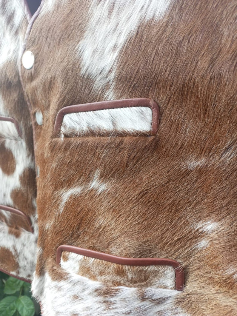 Pu&ograve; includere: Un giubbotto in pelle di mucca marrone e bianco con rifiniture in pelle marrone e due tasche. Il giubbotto ha una chiusura a bottone.