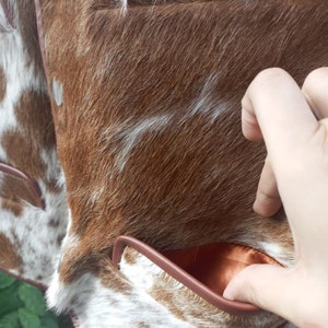 Pu&ograve; includere: Primo piano di una giacca in pelle di mucca marrone e bianca con una tasca in pelle marrone. La tasca &egrave; foderata con un tessuto marrone.