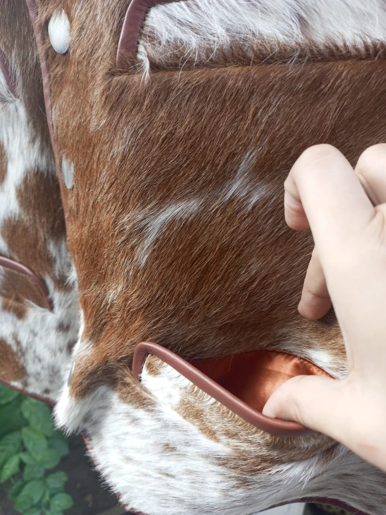 Pu&ograve; includere: Primo piano di una giacca in pelle di mucca marrone e bianca con una tasca in pelle marrone. La tasca &egrave; foderata con un tessuto marrone.