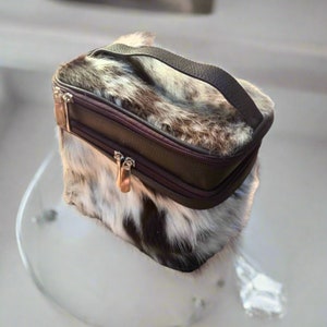 Puede incluir: Una bolsa de maquillaje de cuero marrón y piel de vaca con cierre de cremallera y asa superior.