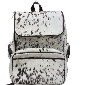 Puede incluir: Una mochila de cuero de vaca blanca y marrón con un bolsillo frontal con cremallera y correas ajustables.