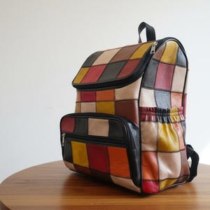Peut inclure: Sac &agrave; dos en cuir patchwork color&eacute;, avec une poign&eacute;e sup&eacute;rieure noire, une fermeture &eacute;clair et des poches. Le sac pr&eacute;sente une vari&eacute;t&eacute; de couleurs, dont le rouge, le jaune, le marron et le beige. Le sac est pos&eacute; sur une table en bois.