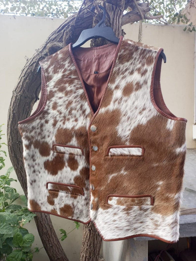 Pu&ograve; includere: Un gilet in pelle di mucca marrone e bianco con chiusura a bottone e due tasche su ogni lato. Il gilet ha un bordo in pelle marrone.