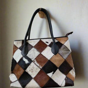 Op de afbeelding: Een bruine en witte patchwork tote bag met zwarte leren handvatten en accenten. De tas is gemaakt van verschillende stukken rundleer, elk met een uniek patroon.