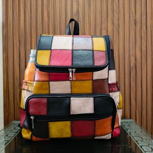 Peut inclure: Un sac &agrave; dos en cuir patchwork multicolore avec une poign&eacute;e noire et des fermetures &eacute;clair. Le sac pr&eacute;sente des carr&eacute;s de couleurs vives : rouge, jaune, noir et blanc. Une gourde est visible dans la poche lat&eacute;rale.