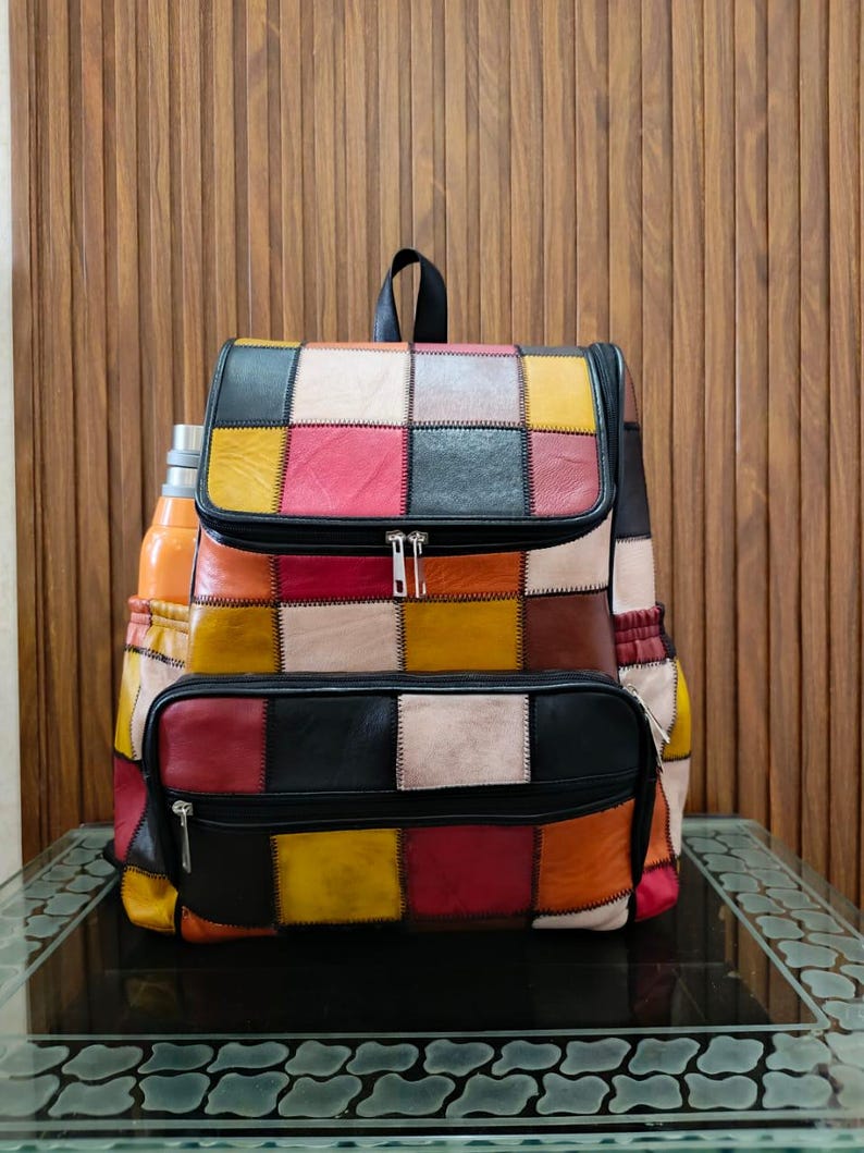 Peut inclure: Un sac &agrave; dos en cuir patchwork multicolore avec une poign&eacute;e noire et des fermetures &eacute;clair. Le sac pr&eacute;sente des carr&eacute;s de couleurs vives : rouge, jaune, noir et blanc. Une gourde est visible dans la poche lat&eacute;rale.