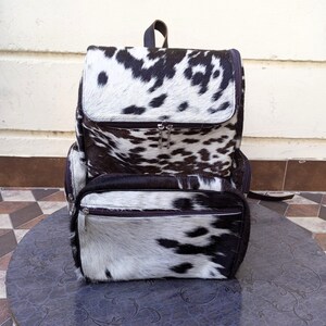 May include: A black and white cowhide leather backpack with two large zippered compartments. The backpack has a brown leather strap and a brown leather trim.