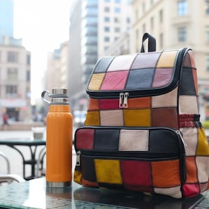 Peut inclure: Un sac &agrave; dos en cuir patchwork multicolore avec une poign&eacute;e sup&eacute;rieure et deux compartiments zipp&eacute;s. Une gourde isotherme orange avec un couvercle argent&eacute; est pos&eacute;e &agrave; c&ocirc;t&eacute; du sac &agrave; dos sur une table en verre.