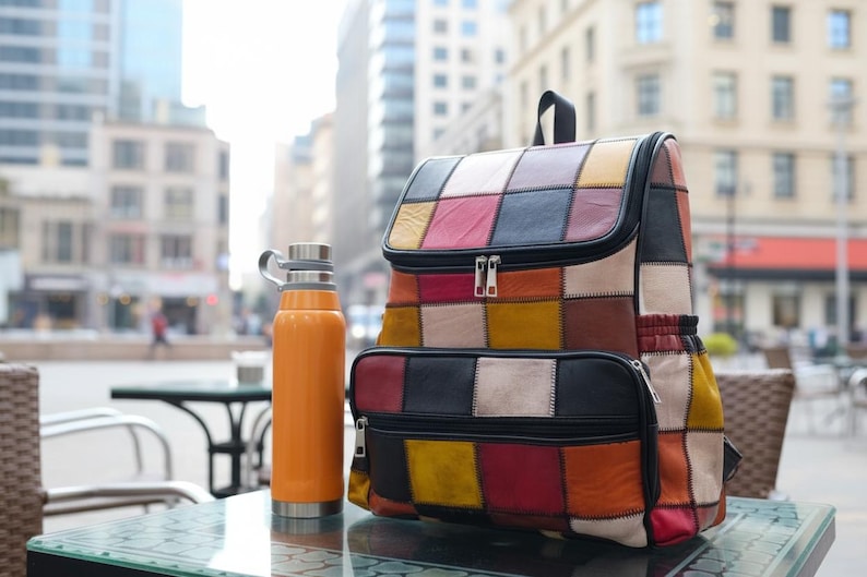 Peut inclure: Un sac &agrave; dos en cuir patchwork multicolore avec une poign&eacute;e sup&eacute;rieure et deux compartiments zipp&eacute;s. Une gourde isotherme orange avec un couvercle argent&eacute; est pos&eacute;e &agrave; c&ocirc;t&eacute; du sac &agrave; dos sur une table en verre.