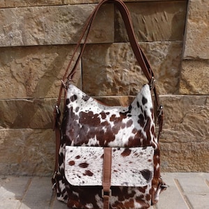 May include: A brown and white cowhide leather shoulder bag with a front pocket and brown leather straps. The bag has a flap pocket with a leather strap closure. The bag is set against a textured stone wall.