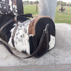 Puede incluir: Bolsos de lona hechos de piel de vaca, con una mezcla de patrones de piel negros, blancos y marrones. Cada bolso tiene una correa de cuero marr&oacute;n y cierre de cremallera. Los bolsos son de forma cil&iacute;ndrica.