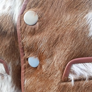 Pu&ograve; includere: Primo piano di un gilet in pelle di mucca marrone e bianco con tre bottoni argentati. Il gilet ha un bordo in pelle marrone.