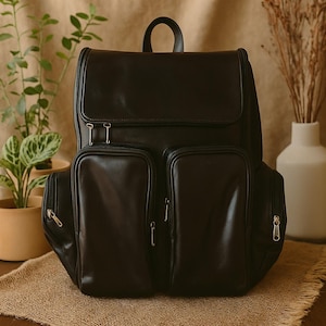 May include: A black leather backpack with multiple zippered compartments. The backpack has a top handle and a flap closure. The bag is sitting on a wooden table with a burlap cloth. Plants and a vase are in the background.