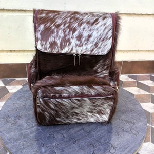 May include: A brown and white cowhide backpack with two zippered compartments. The backpack has a brown leather trim and a white zipper pull.