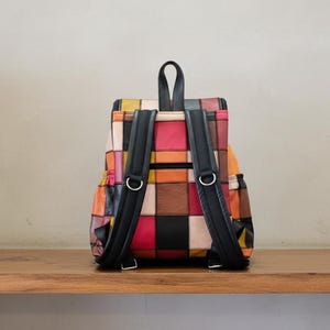 Peut inclure: Sac &agrave; dos patchwork color&eacute; avec des bretelles noires et des anneaux m&eacute;talliques argent&eacute;s. Le sac &agrave; dos pr&eacute;sente une vari&eacute;t&eacute; de couleurs, dont le rouge, l'orange, le jaune et le noir. Pos&eacute; sur une surface en bois.