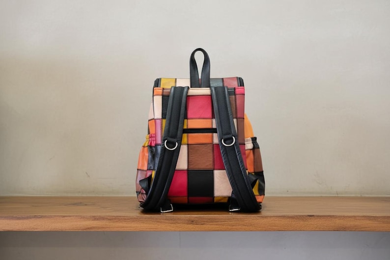 Peut inclure: Sac &agrave; dos patchwork color&eacute; avec des bretelles noires et des anneaux m&eacute;talliques argent&eacute;s. Le sac &agrave; dos pr&eacute;sente une vari&eacute;t&eacute; de couleurs, dont le rouge, l'orange, le jaune et le noir. Pos&eacute; sur une surface en bois.