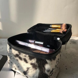 May include: A rectangular makeup bag with a black and white cow print pattern. The bag features a black zipper closure and a black interior lining. Inside, the bag contains several makeup brushes with gold ferrules and a palette with multiple shades.